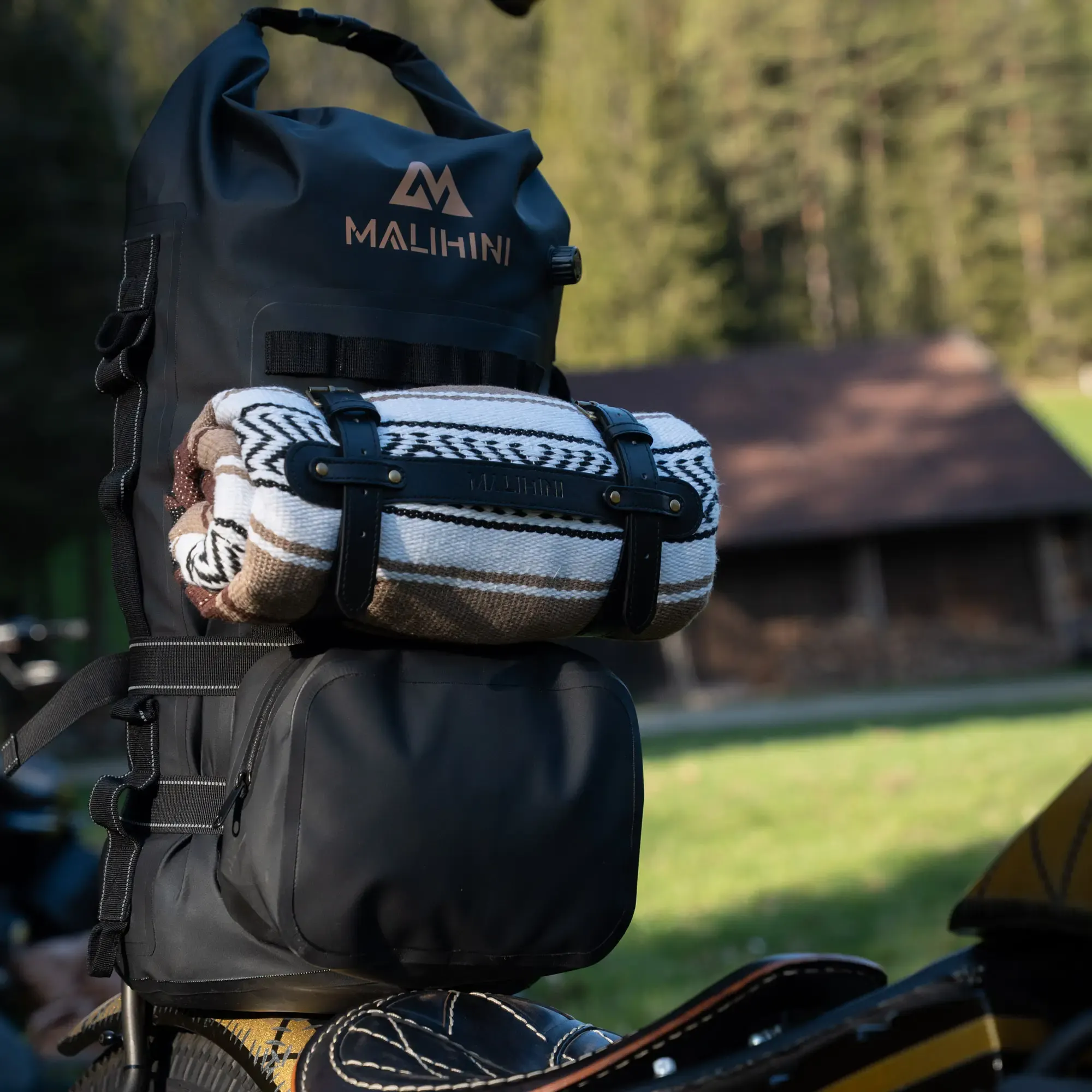 Malibag 46L Drybag am Motorrad, ideal für Camping. Schwarze Tasche mit zusätzlicher Decke befestigt, im Hintergrund eine Holzhütte und Wälder.